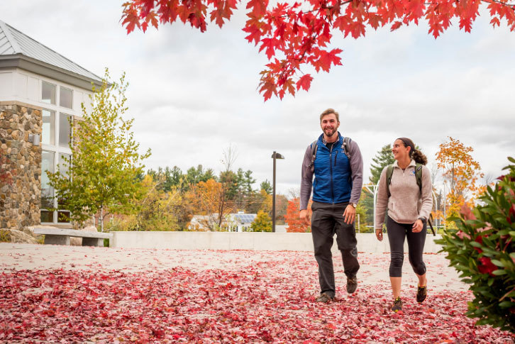 Two students walking down a sidewalk covered with red leaves.