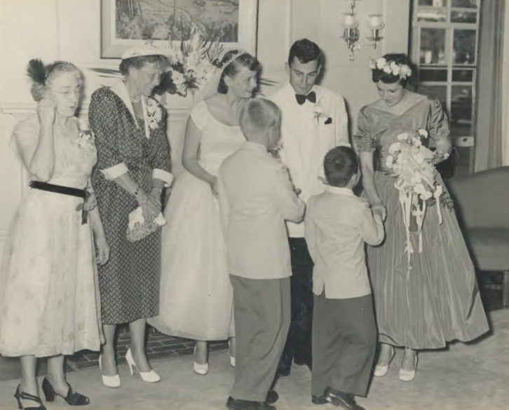 A line-up of the bride, groom, maid of honor and their mothers. The backs of two small boys are seen as they are facing the newly wedding couple and shaking hands with them.